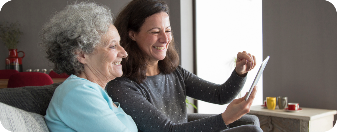 Two women sit on a couch and smile while discussing the advantages of home care compared to the disadvantages of an aged care facility. 