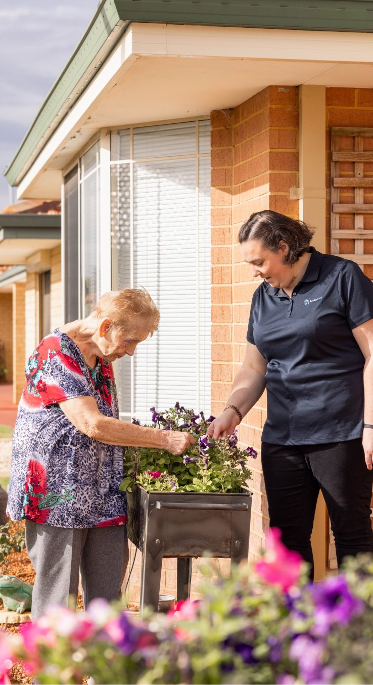 Care recipient enjoys home care gardening activities while working on a flower bed outside with assistance from her caregiver.
