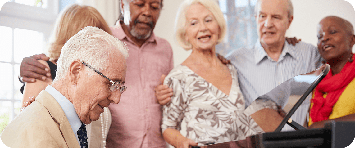 A photo of older people gathered around a piano and joyfully singing together.